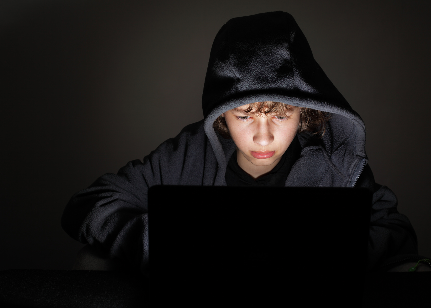 A young person wearing a hood intently looks at a laptop screen, which illuminates their face in a dark room.