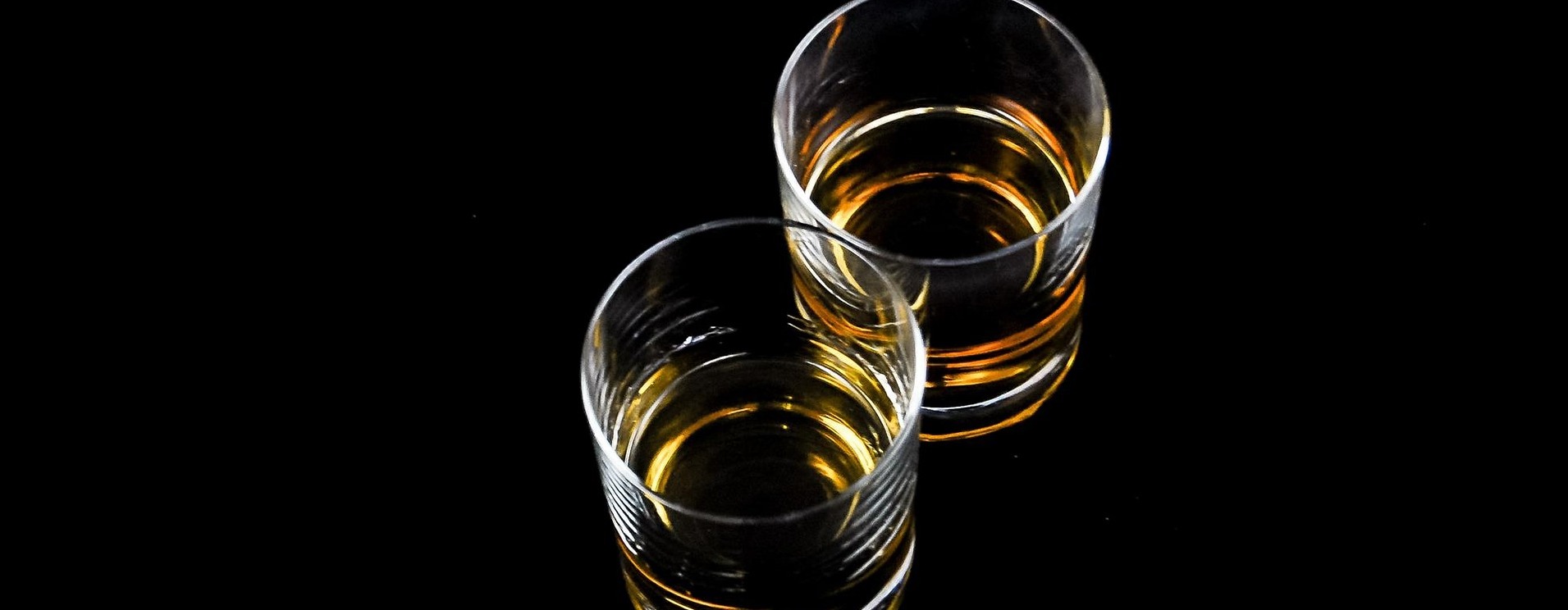 Two glasses of whiskey on a reflective black surface, highlighted by a soft light that accentuates their amber contents.