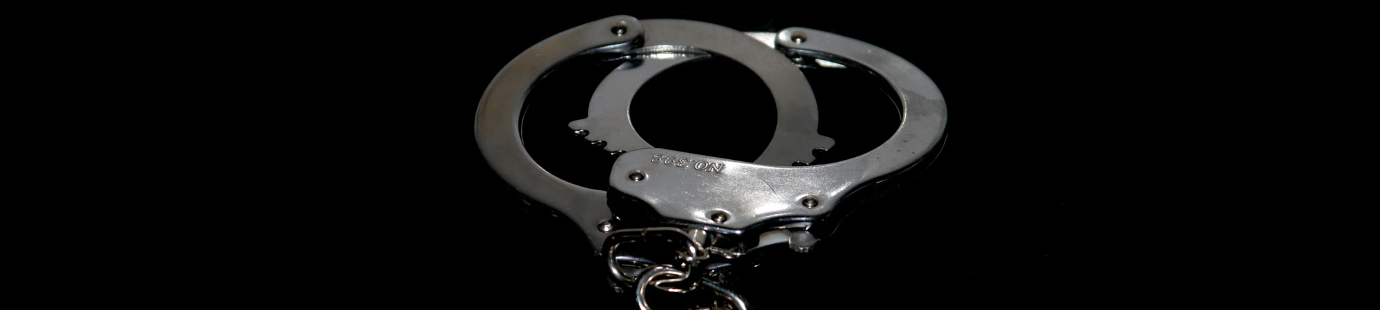 A pair of open, silver handcuffs laying on a reflective black surface under a dim light, creating a dramatic effect with sharp contrasts.