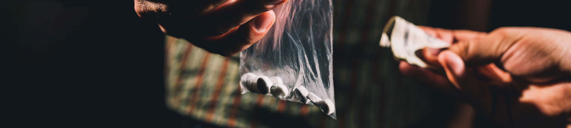 A close-up image of a person holding a small zip-lock bag with pills while another hand holds cash, focusing on an apparent transaction in dim lighting.