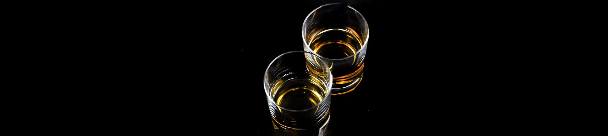 Two glasses of whiskey with amber liquid, reflected on a shiny black surface against a dark background, creating a symmetrical visual.