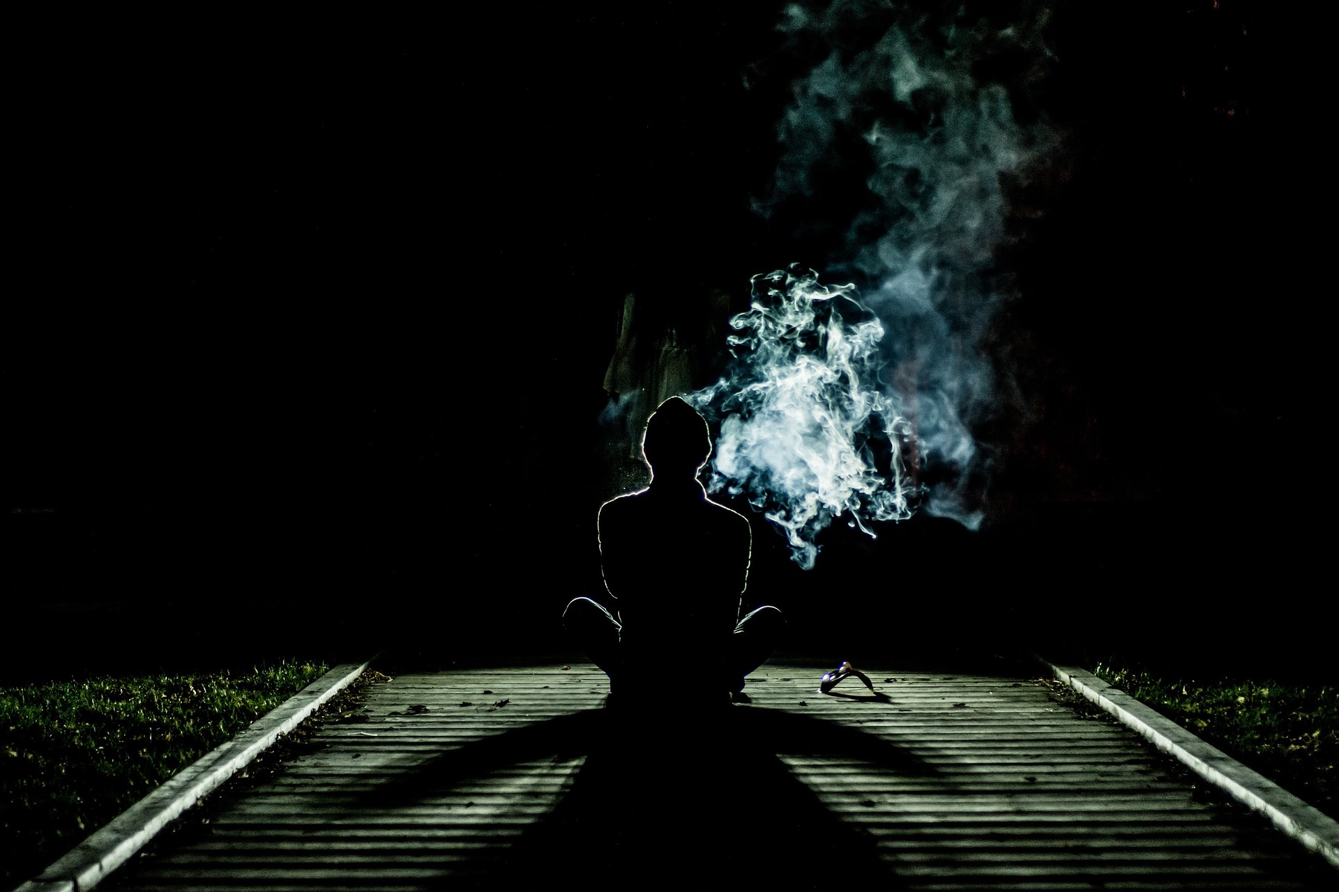 A person sits cross-legged on a wooden dock at night, silhouetted against the dark, with smoke from a controlled substance rising artistically into the air around them. A subtle greenish light