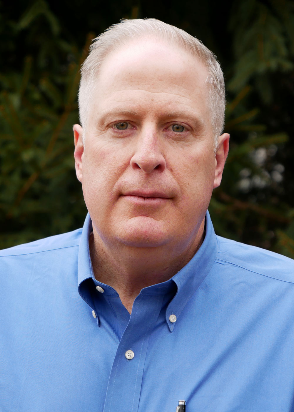 A headshot of a middle-aged man with short blond hair, wearing a blue button-down shirt, posing against a background of blurred green foliage.