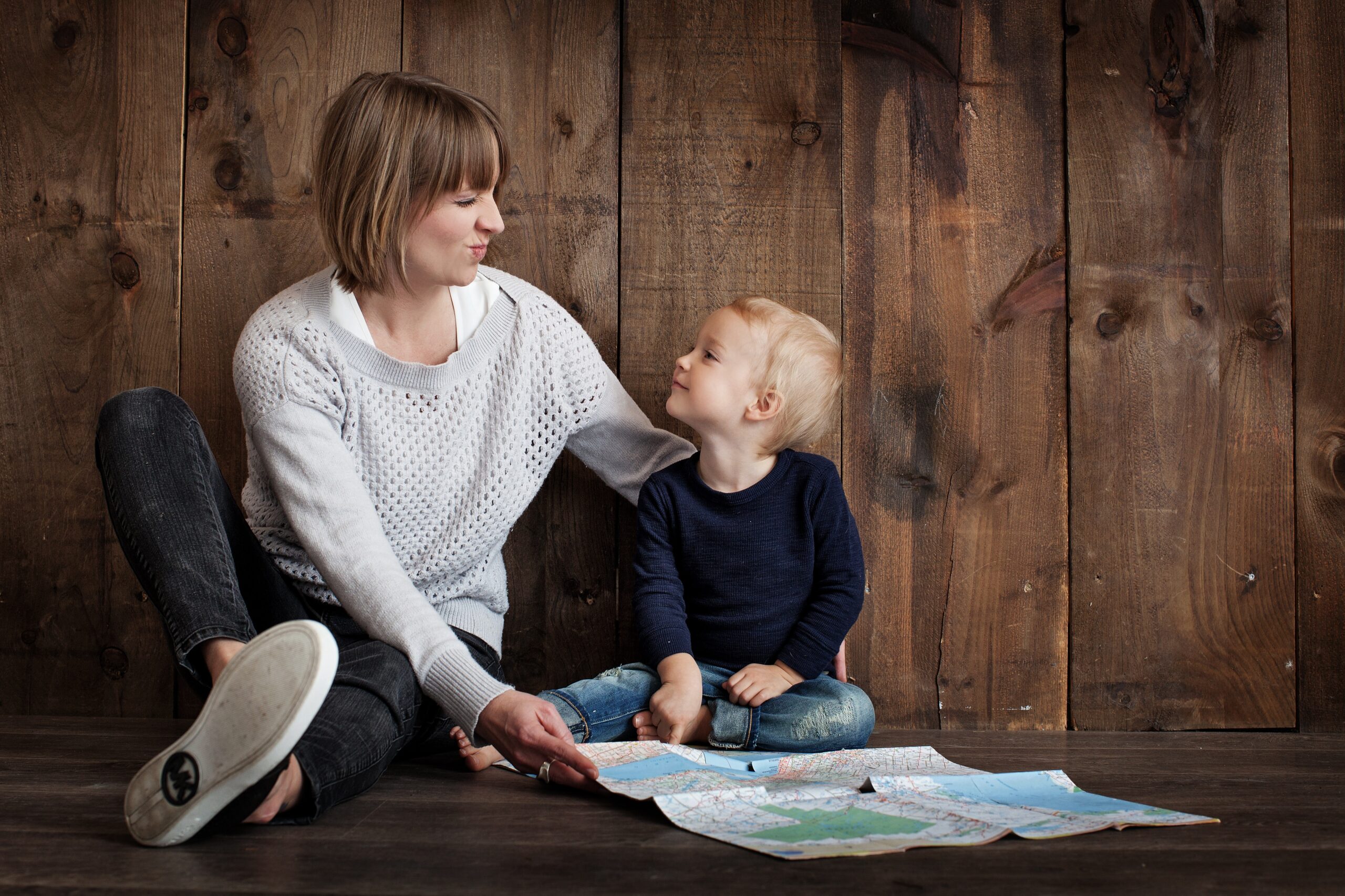 A mother and her young son sit on a wooden floor, gazing at each other with smiles while surrounded by spread out maps, engaged in permanency planning together.