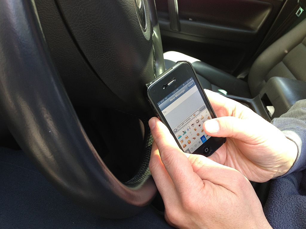 A person using a smartphone with a qwerty keyboard interface while sitting in the driver's seat of a car in New Jersey, holding the phone near the steering wheel with both hands.