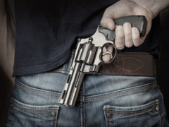 A person in New Jersey wearing denim jeans holds a silver revolver tucked into their waistband, gripping the handle with one hand.