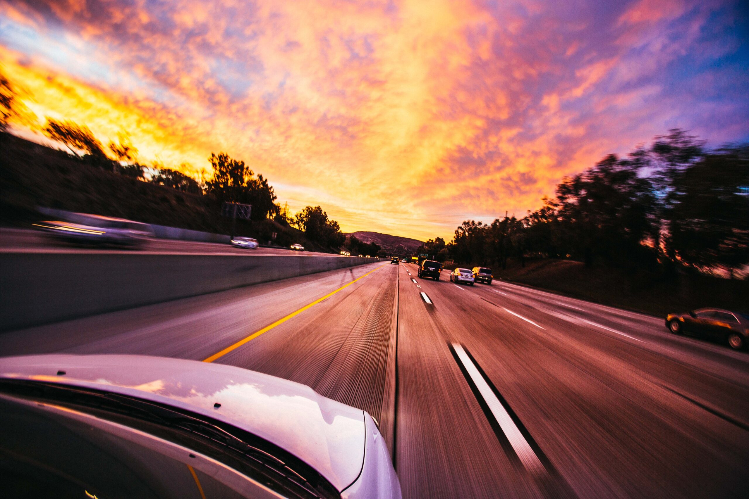 A dynamic view from a moving vehicle on a highway at sunset, with vibrant orange and pink clouds in the sky and blurred motion of the road and surrounding cars, reminiscent of the consequences of speeding.