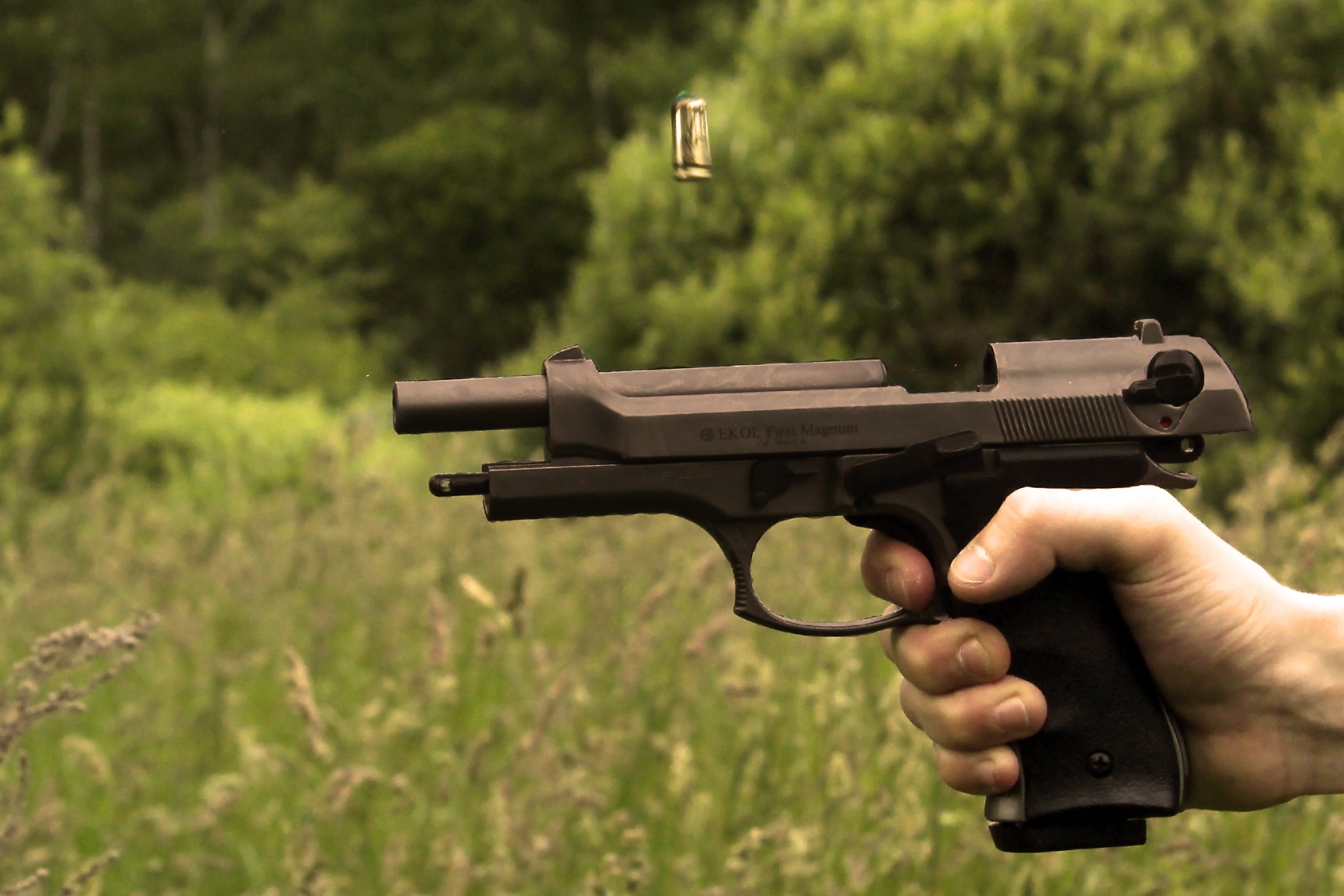 A close-up of a hand illegally possessing a black pistol with a bullet casing ejecting midair, against a blurred green forest background.