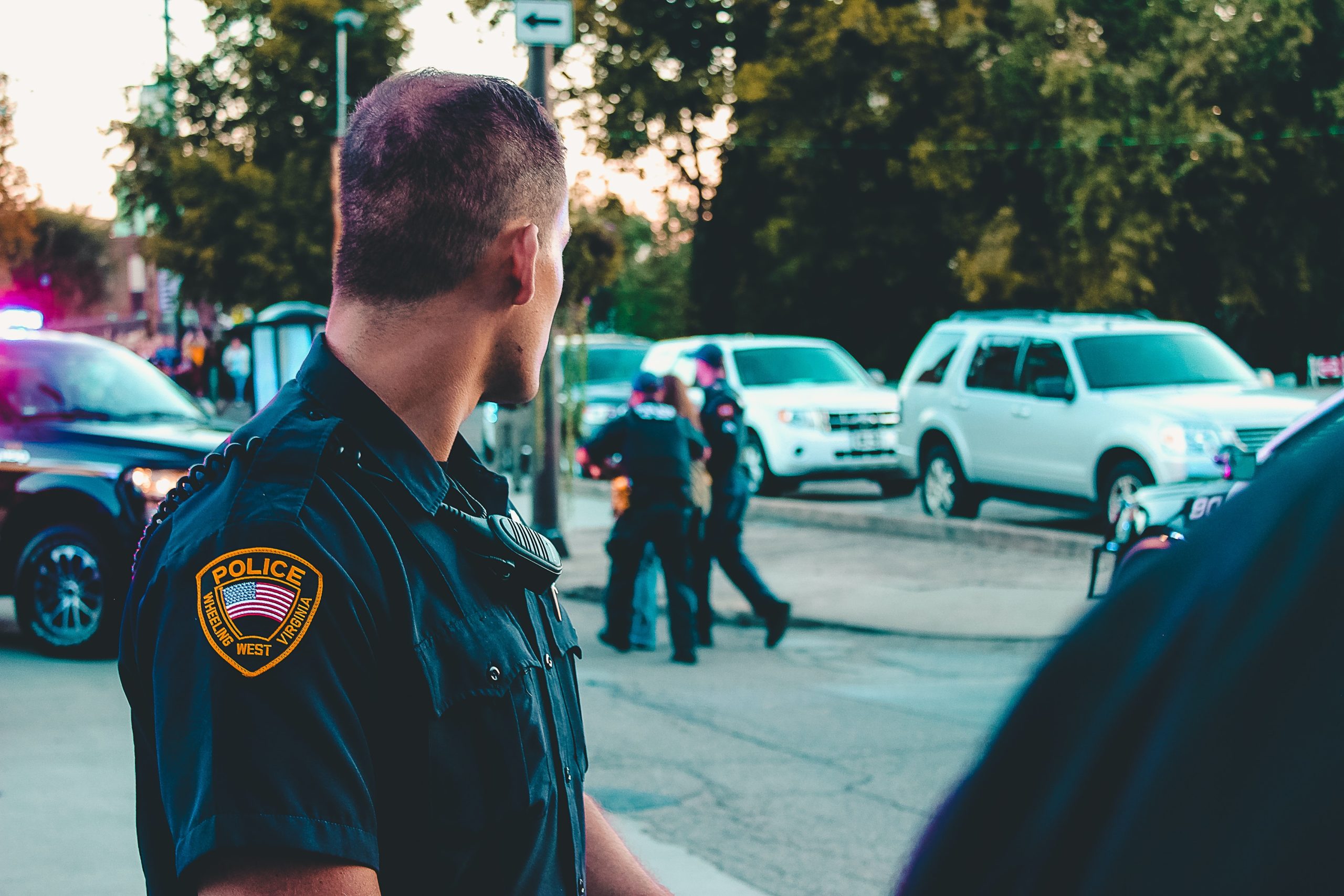 A police officer in uniform, viewed from the side, watches over a busy street scene where other officers interact with the public under a zero-tolerance policy, with police vehicles and civilian cars in the background