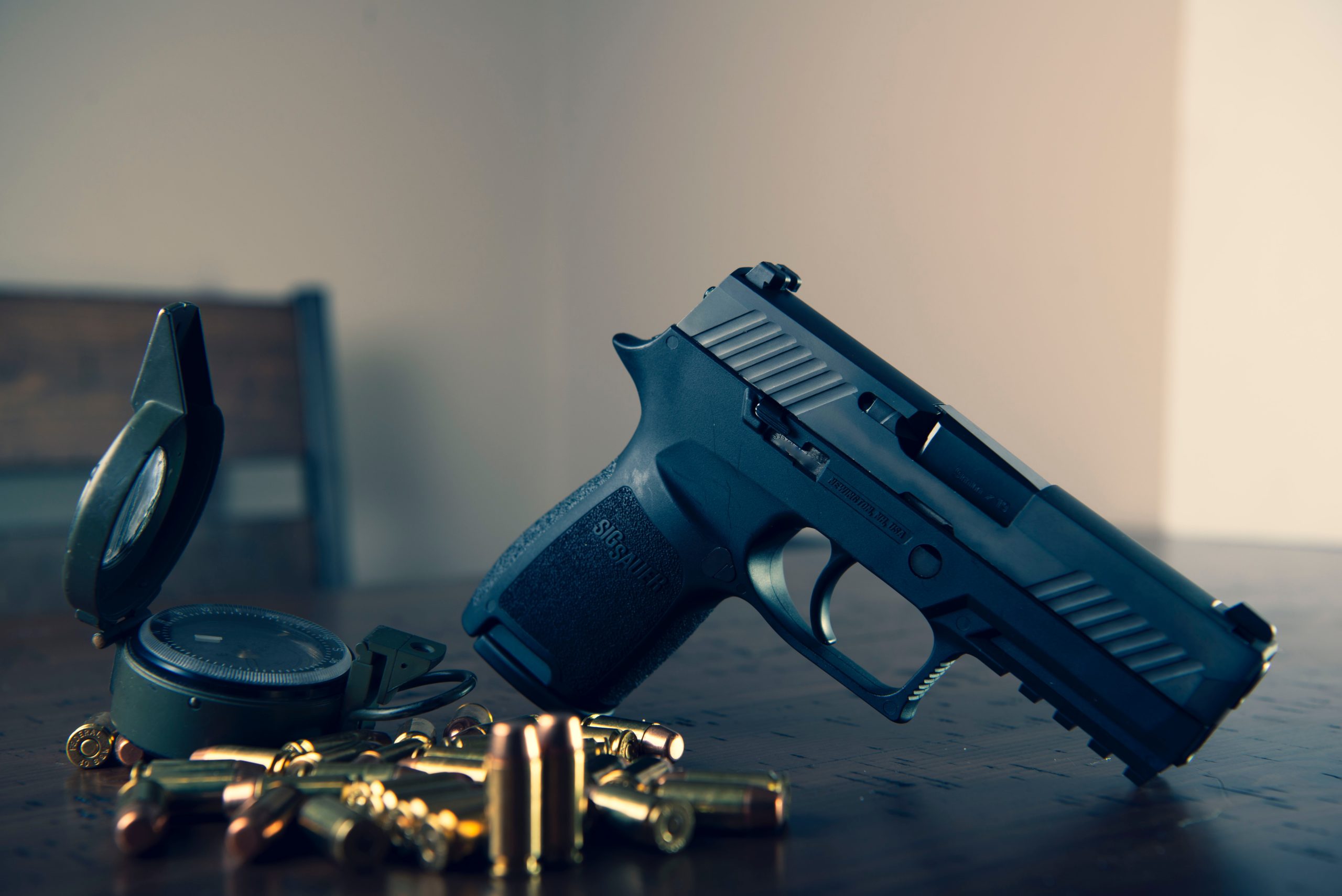 A handgun lies on a wooden surface with scattered bullets and a compass next to it, suggesting themes of travel and security. The setting is dimly lit, emphasizing a dramatic atmosphere potentially linked to gun laws