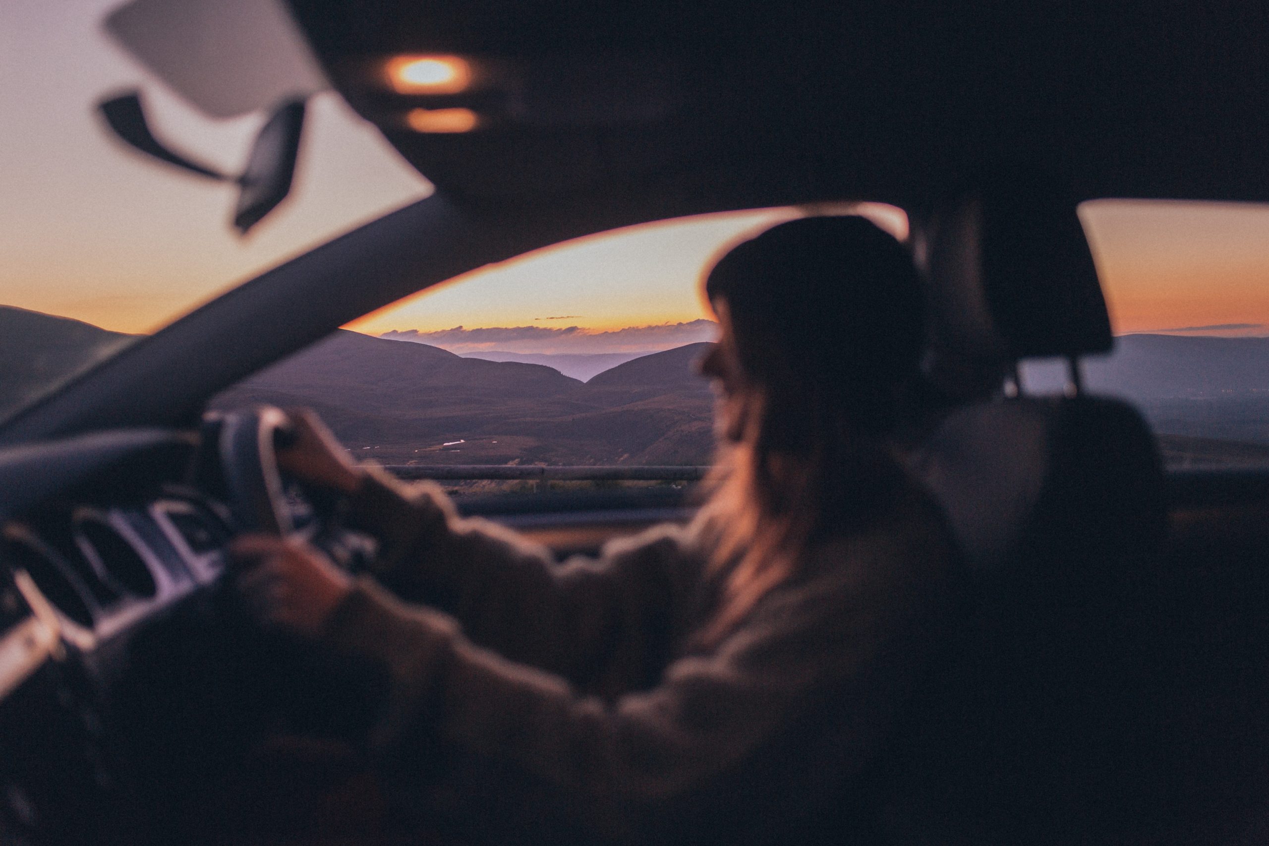 A woman driving a car during sunset in New Jersey, with a scenic view of layered mountains and a soft orange sky visible through the windshield.