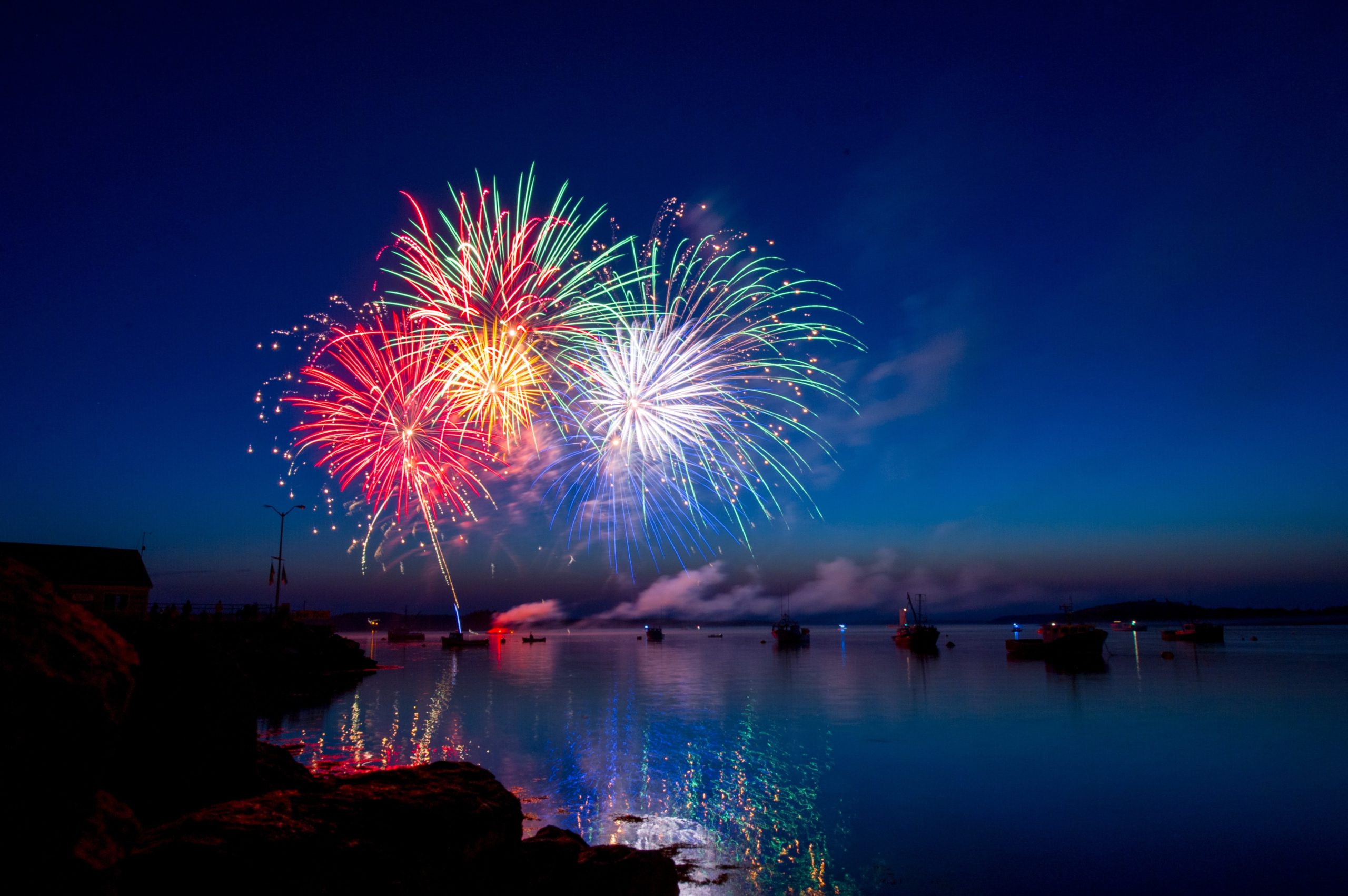 Colorful fireworks bursting over a calm bay at dusk, with boats and a rocky shoreline visible. Reflections of the vibrant red, green, and white lights shimmer on the water's surface as nearby,