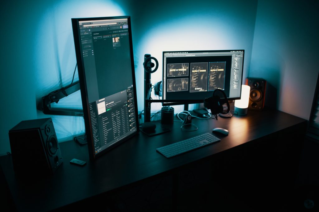 A dimly lit workspace featuring two computer monitors displaying a music production software, a keyboard, headphones, and a speaker, all illuminated by blue ambient lighting and a sticky note warning against identity theft.