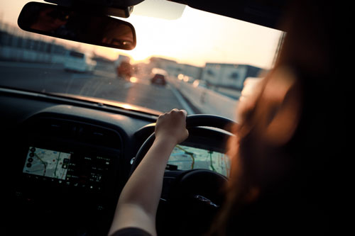 A person making an unsafe lane change while driving a car during sunset, viewed from the backseat, showing one hand on the steering wheel and a GPS navigation screen lit up.