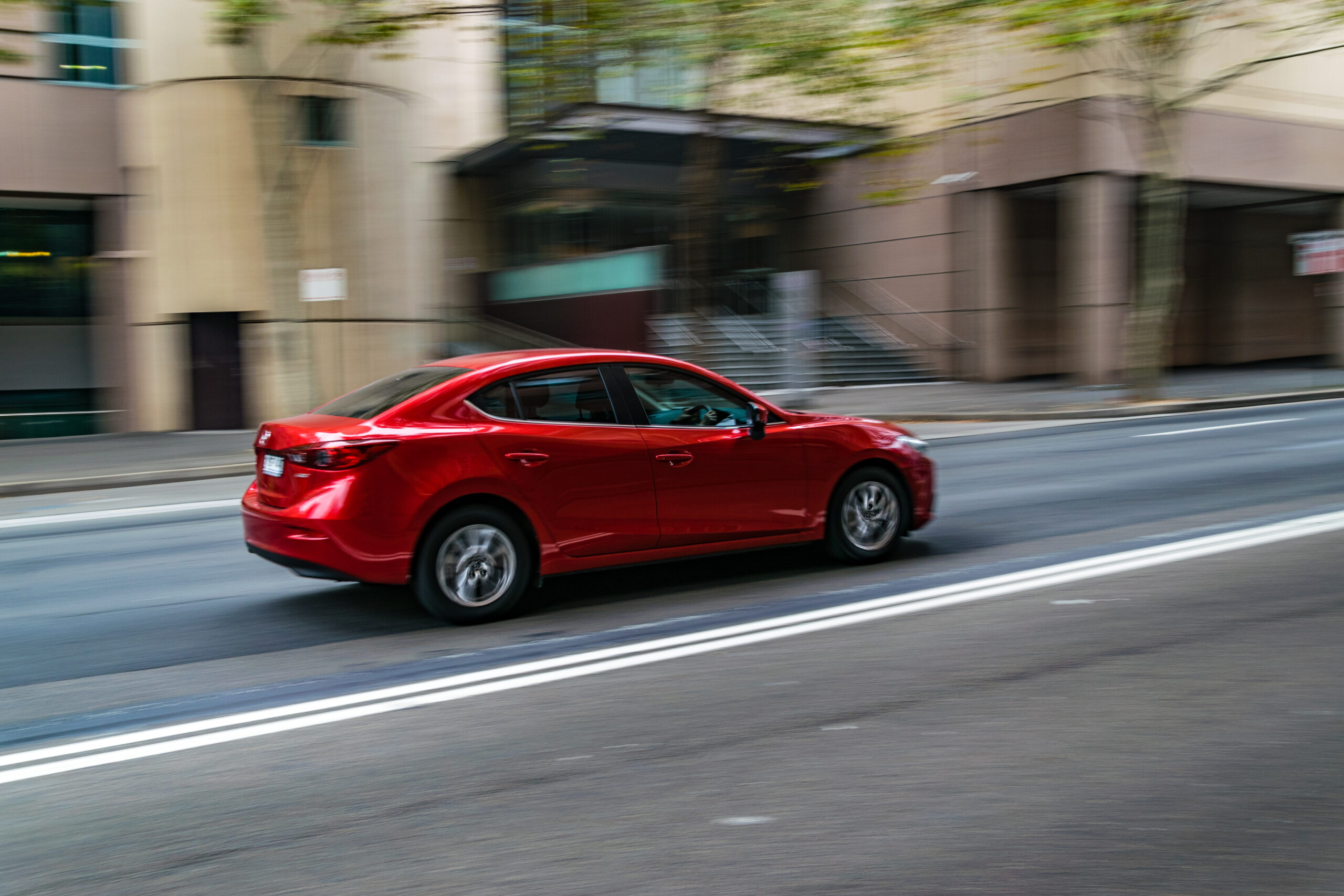 A red sedan in motion, captured with a slow shutter speed to create a motion blur effect, speeding along a city street lined with modern buildings in an unsafe lane.