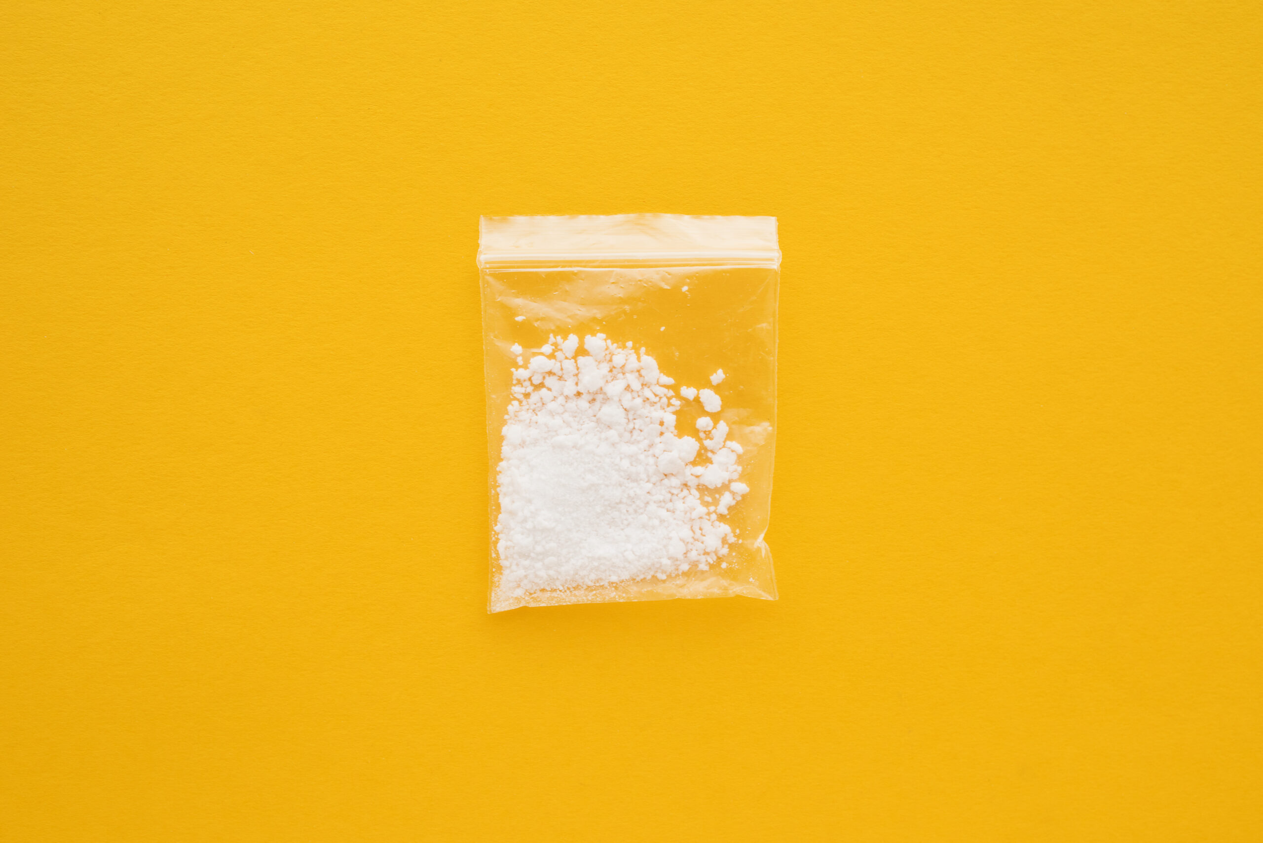 A small clear plastic bag containing cocaine, in the form of white powder and crystalline substances, centered on a vibrant yellow background.