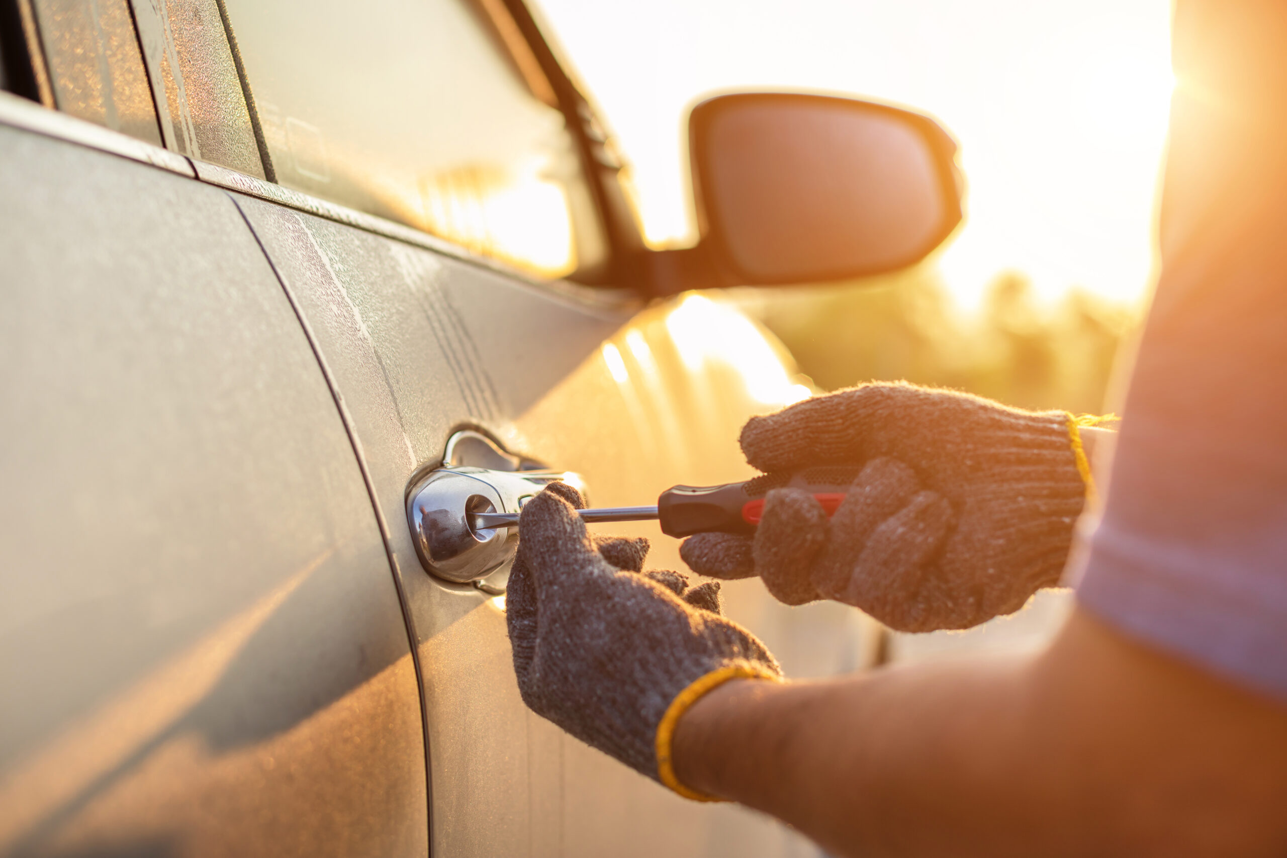 Close-up of a person using a key to unlock a car door, wearing gloves, with sunset light in the background. The focus is on the key entering the lock in an act of theft.