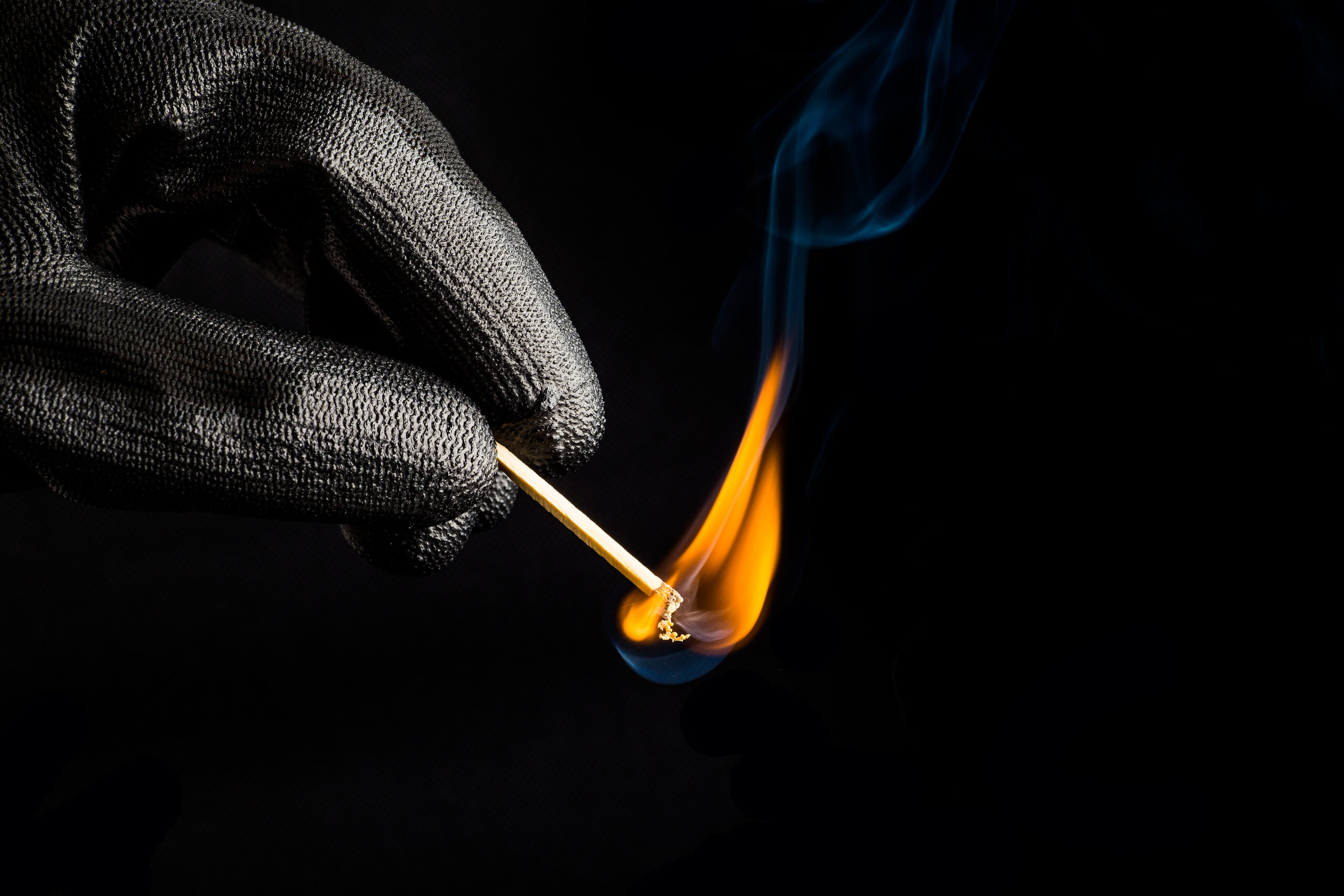 A close-up image of a hand in a silver textured glove holding a lit match, implicated in arson in New Jersey, with a bright orange flame and swirling smoke against a dark background.
