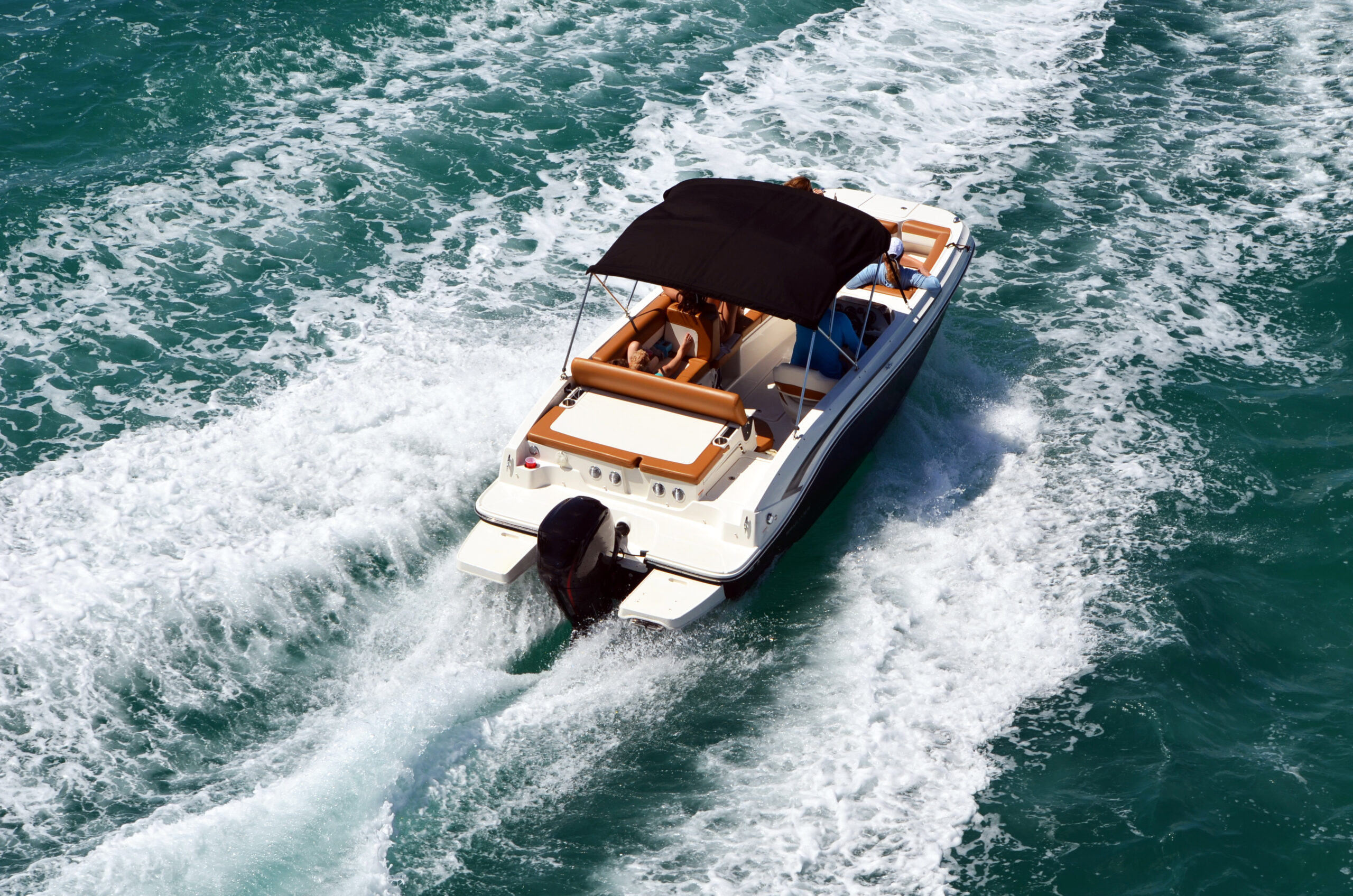 A white motorboat boating over turquoise waters with two people shaded by a black umbrella, viewed from above.