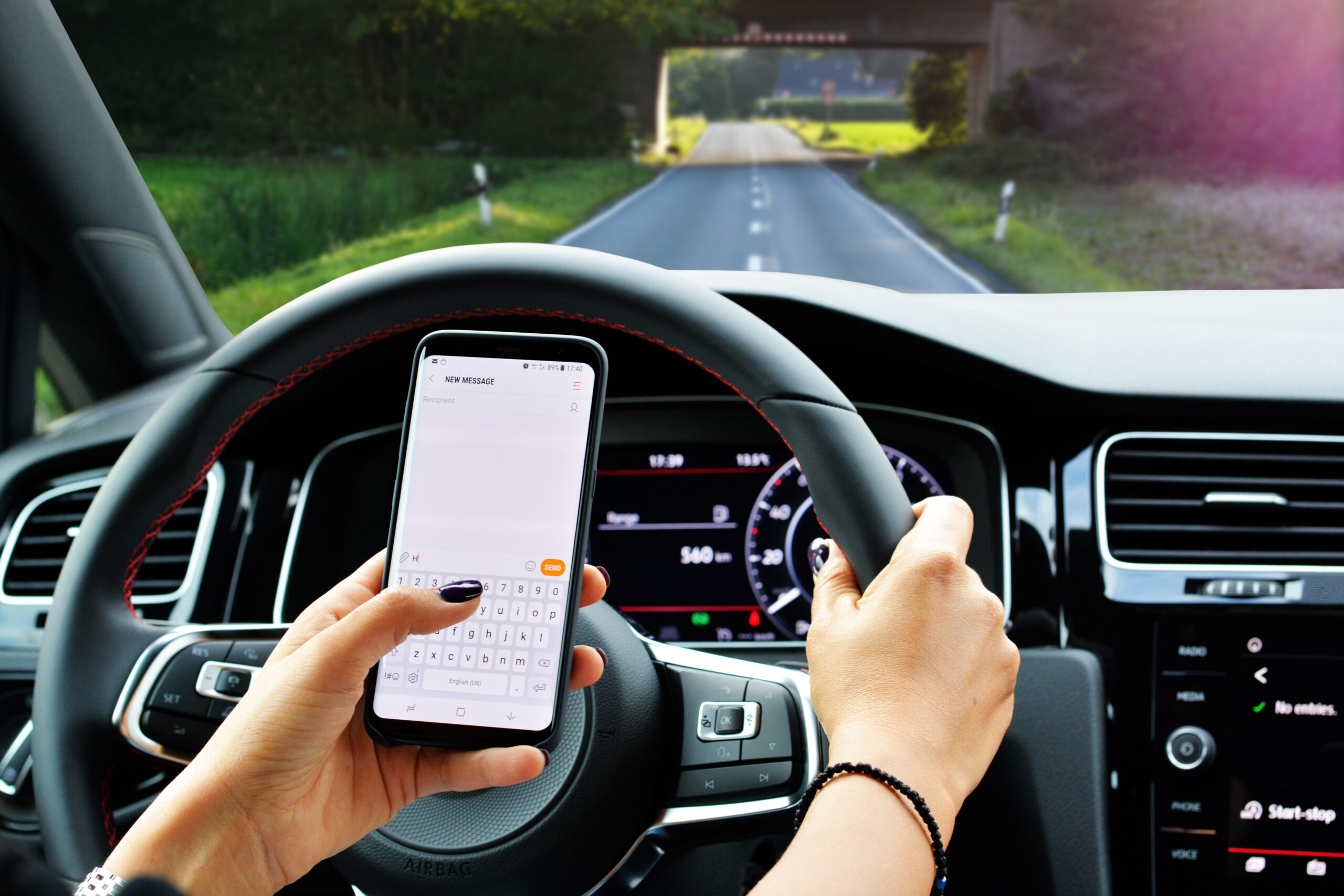 Driver's view inside a car, holding a cell phone and texting while driving on a tree-lined road during daytime, highlighting dangerous behavior.