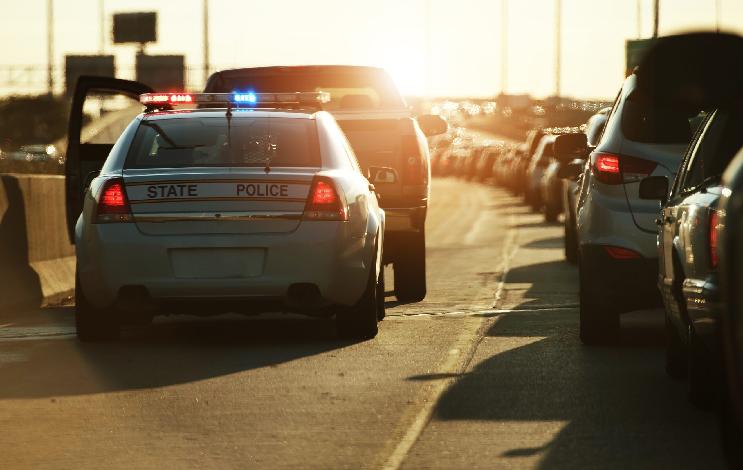 An out-of-state police car with flashing lights stopped on a busy highway during sunset, causing a slight traffic delay.