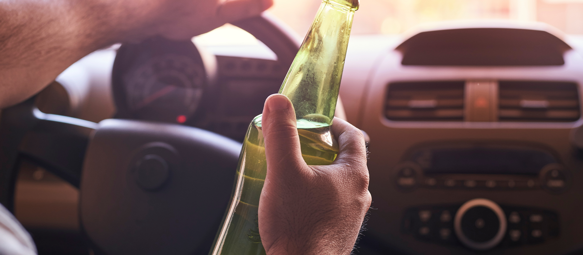 A driver inside a car holding a beer bottle, emphasizing the dangers of drinking and driving, with dashboard visible in the background. sunlight filters through the windshield.