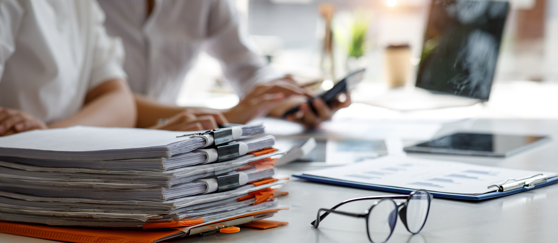 A professional working at a desk with stacks of documents, glasses, and digital devices, focusing on paperwork in a bright office setting.