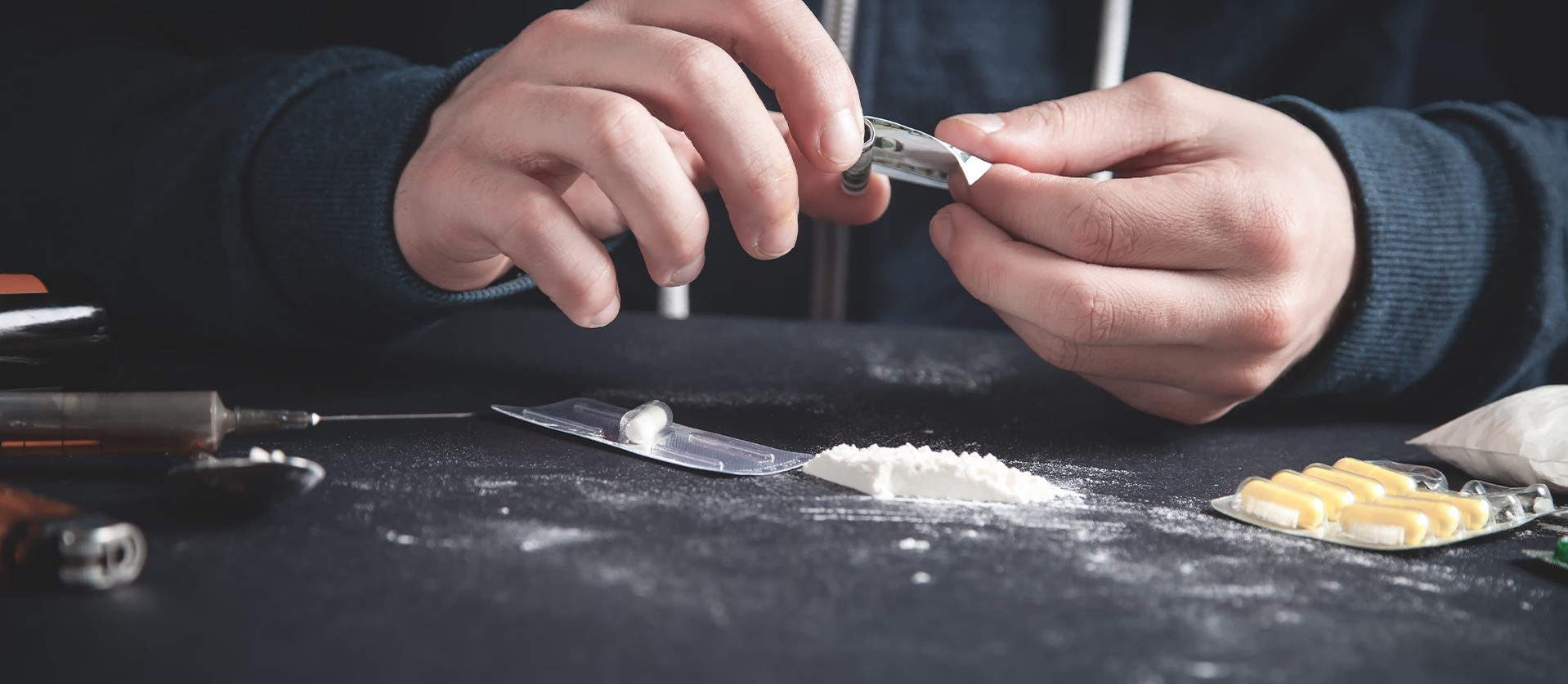 Person handling drugs with tools on a dark surface, including a spoon with powder, pills, and syringes, indicating substance abuse or illegal drug activity.