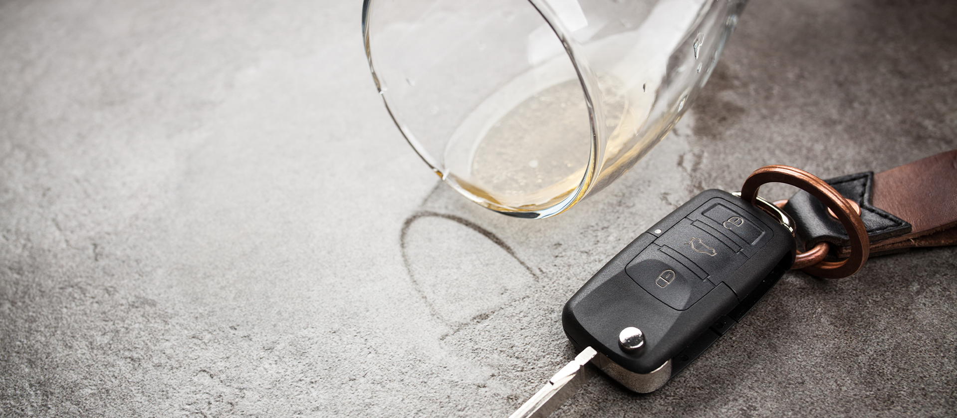 An overturned glass with a small amount of alcohol next to car keys on a concrete surface, suggesting the dangers of drinking and driving.