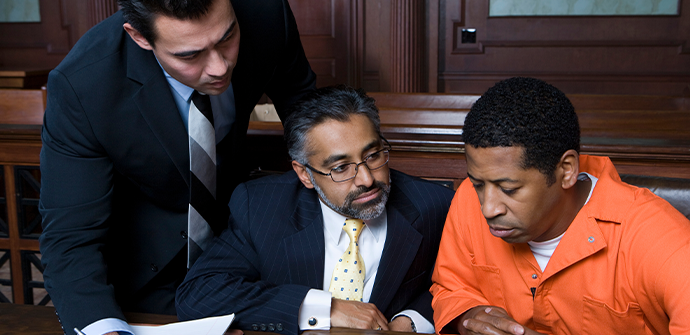 Two Bergen County criminal defense attorneys in suits consulting with a man in an orange jumpsuit, looking at documents inside a courtroom.