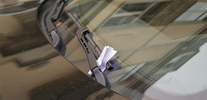 Close-up of a parking ticket placed under a windshield wiper on a car's dusty hood, showing the reflection of nearby objects in the glossy surface.