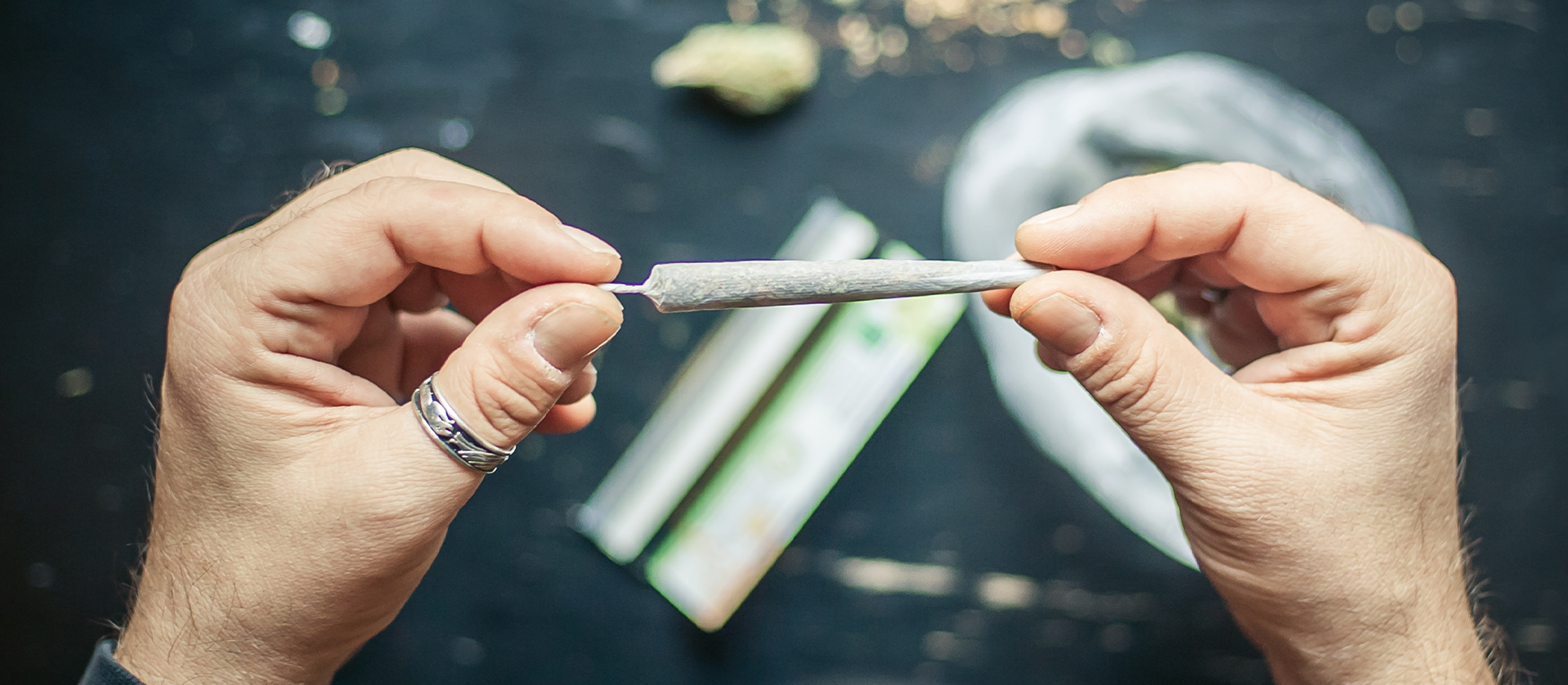Hands rolling a marijuana joint over a black surface, with scattered cannabis leaves and rolling papers visible.