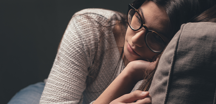 A woman wearing glasses and a sweater rests her head on her arm, which is on a cushion, in a thoughtful or tired pose against a dark background, reminiscent of the contemplative mood often seen in