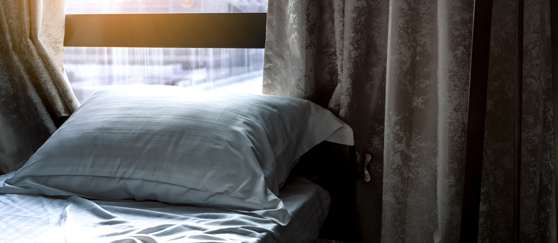 A morning bedroom scene with sunlight filtering through sheer curtains onto a neatly made bed with a fluffy pillow and crisp white sheets.