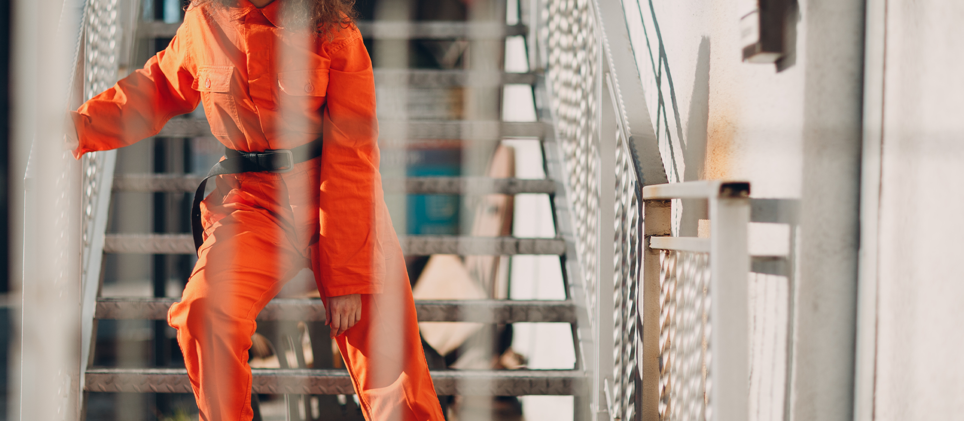 A person in a vibrant orange jumpsuit with a belt poses on a metal staircase, with intricate shadows cast by the sun, emphasizing the urban setting.