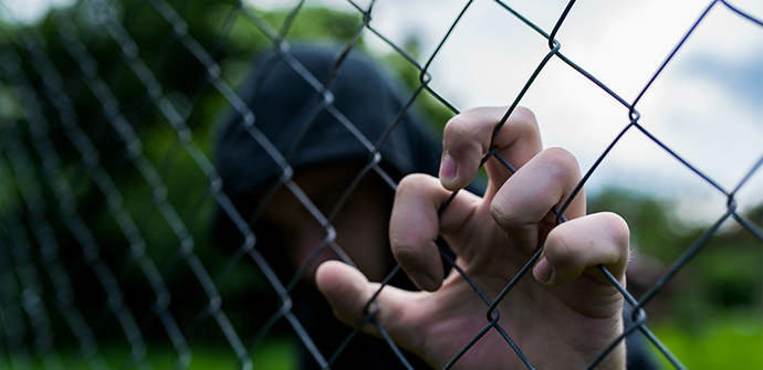 A person in a baseball cap, partially obscured, holds onto a chain-link fence with focus on their hand gripping the metal. Blurred Bergen County background.