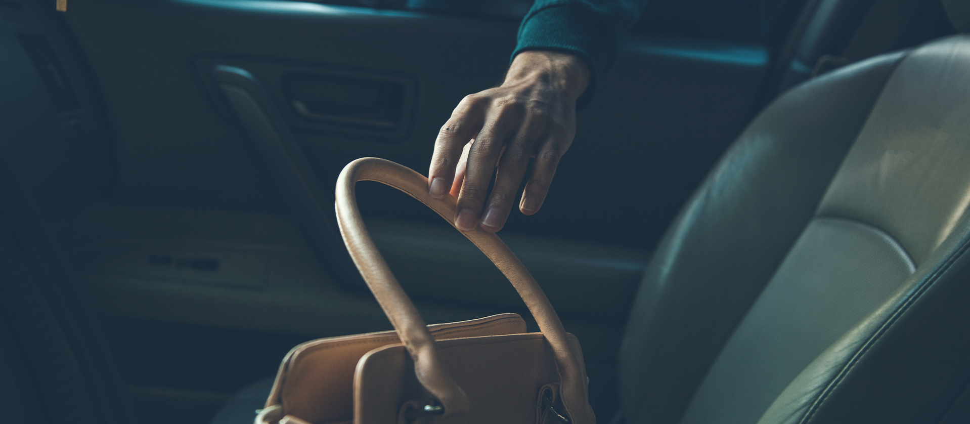 A person reaches into an open car to grab a tan handbag from the passenger seat, with a focus on their hand and the top of the bag.
