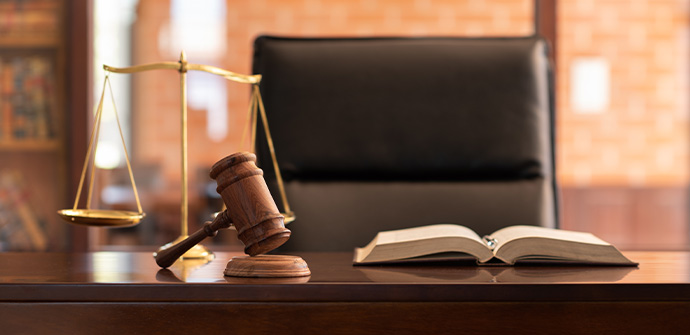 A wooden gavel and a sound block on a desk with an open law book in front and a scale of justice to the left, in an interior design setting with a blurred background.